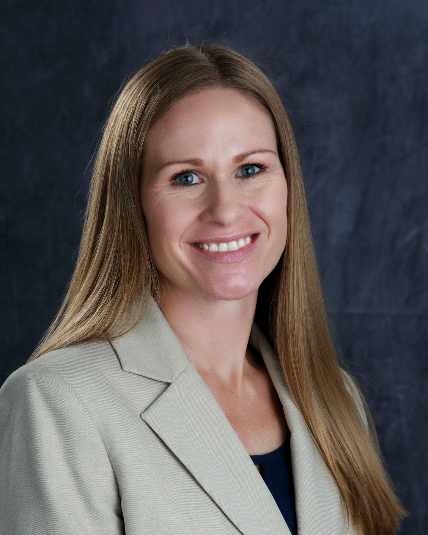 Woman with long blonde hair, smiling, wearing a blazer. Dark blue background.