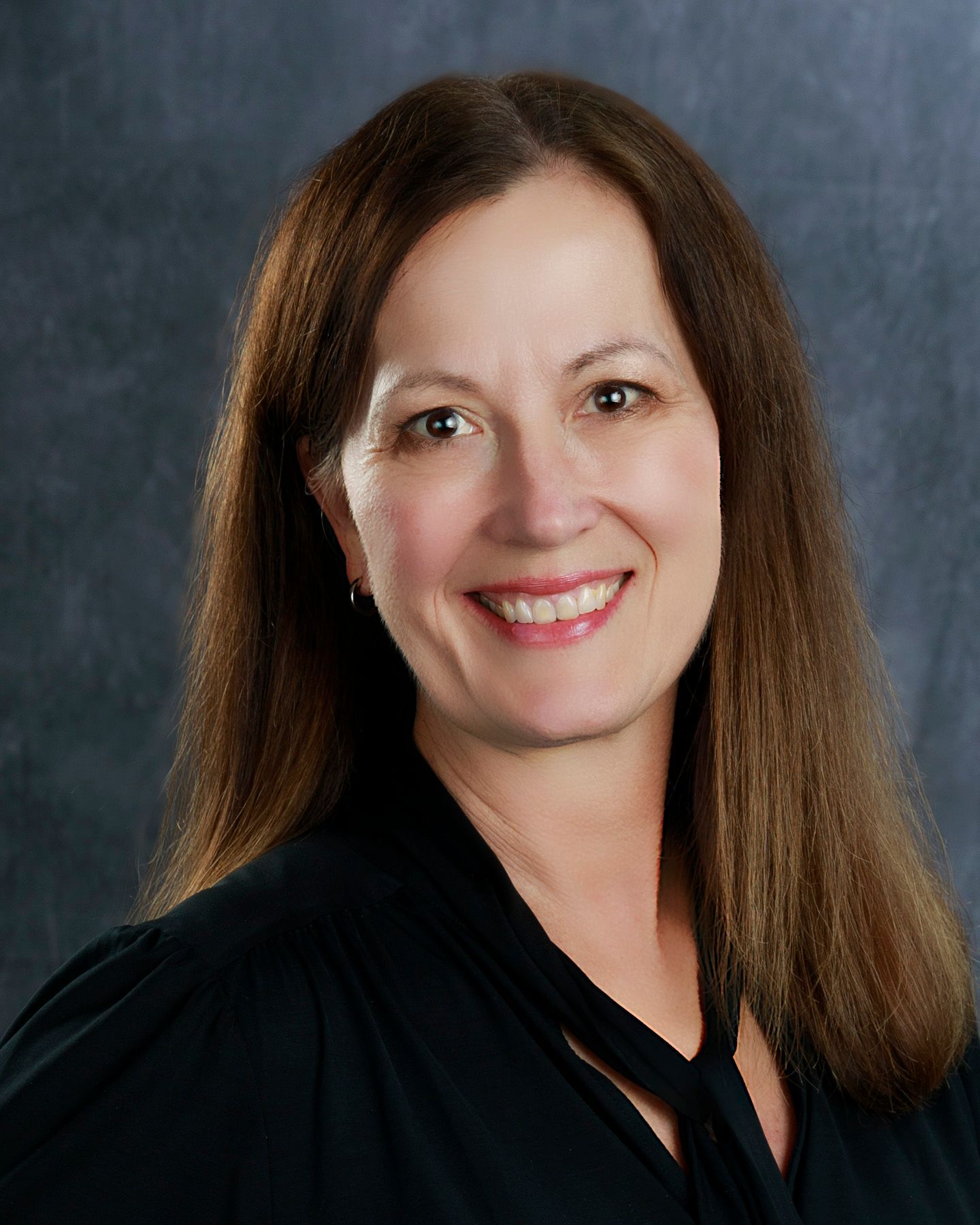 Woman with long brown hair, smiling, wearing a black robe, against a gray background.