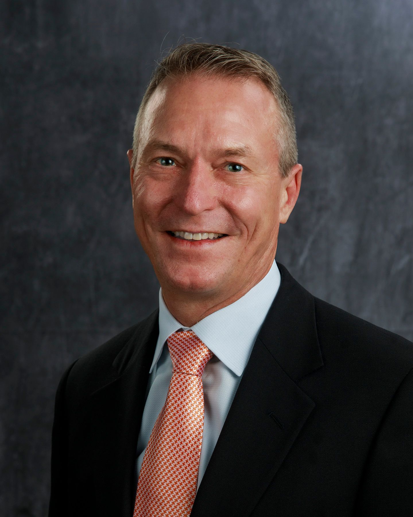 Man in a suit smiles at the camera; peach-colored tie, blue shirt, dark background.