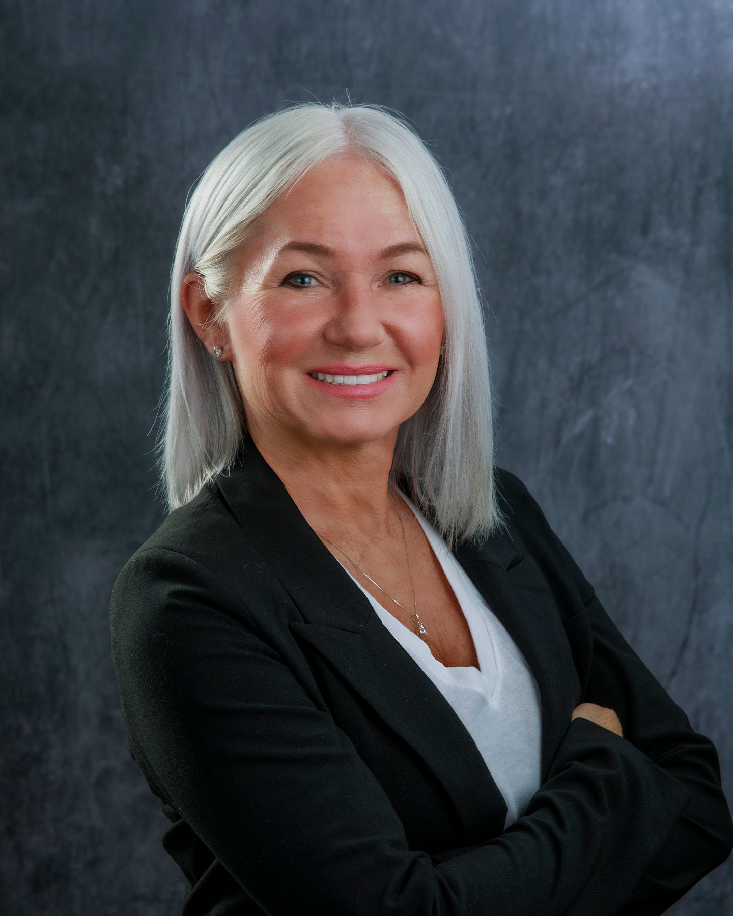 Woman with silver hair, wearing a black blazer and white shirt, smiling with arms crossed.
