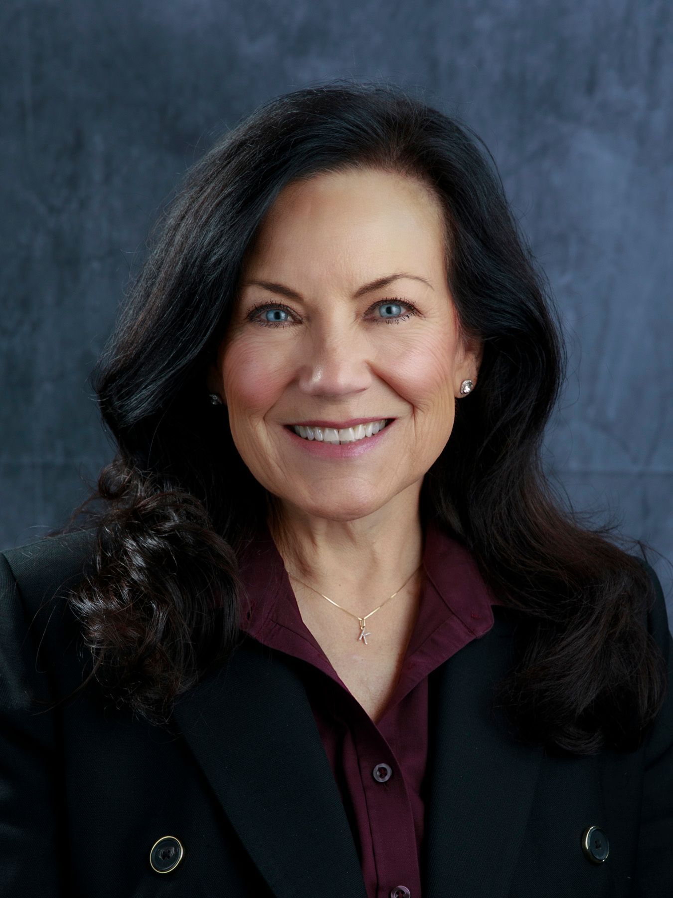 Woman with long dark hair smiles, wearing a burgundy shirt and black blazer against a blurred backdrop.