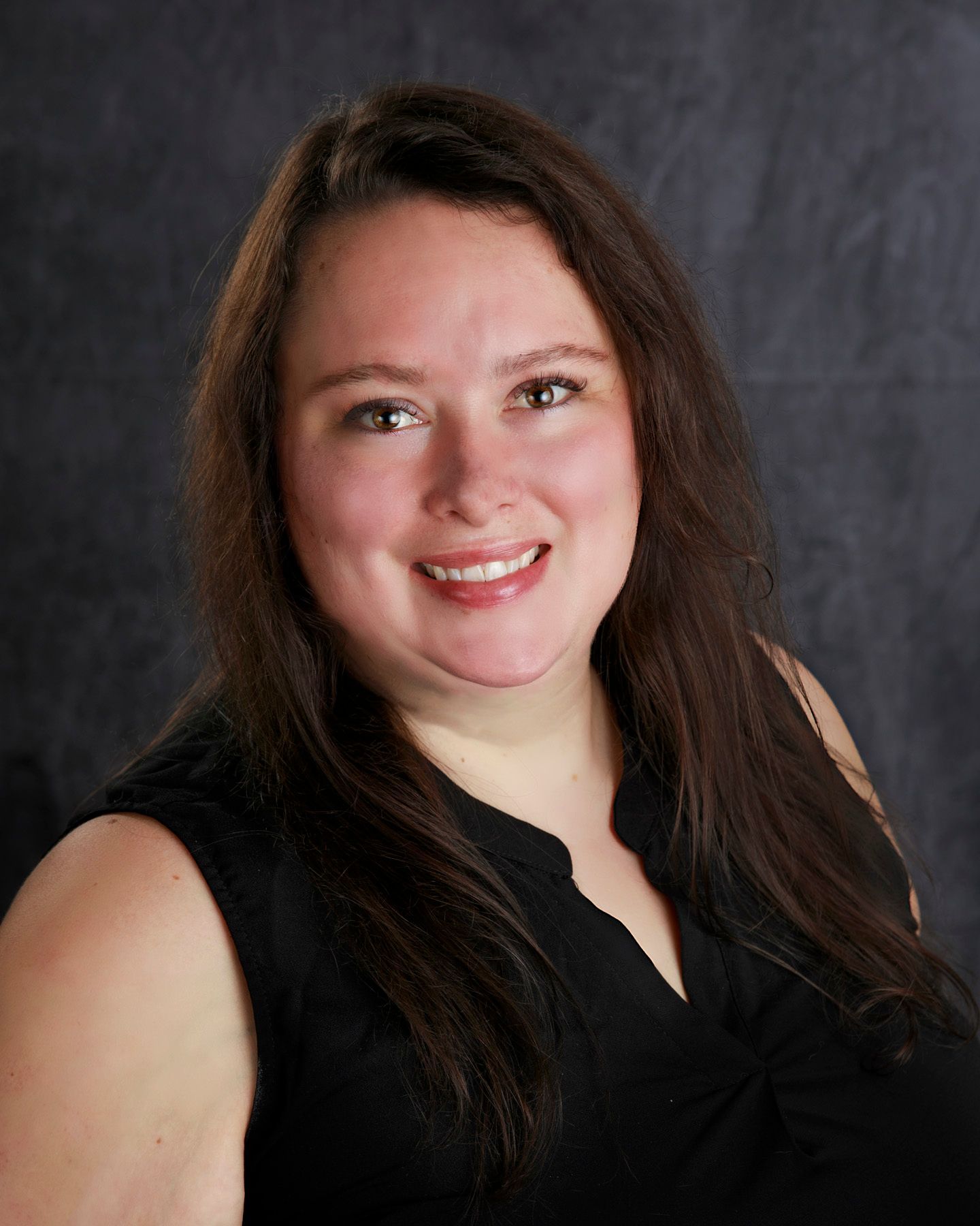 Woman with long brown hair smiles, wearing a black top against a dark gray backdrop.