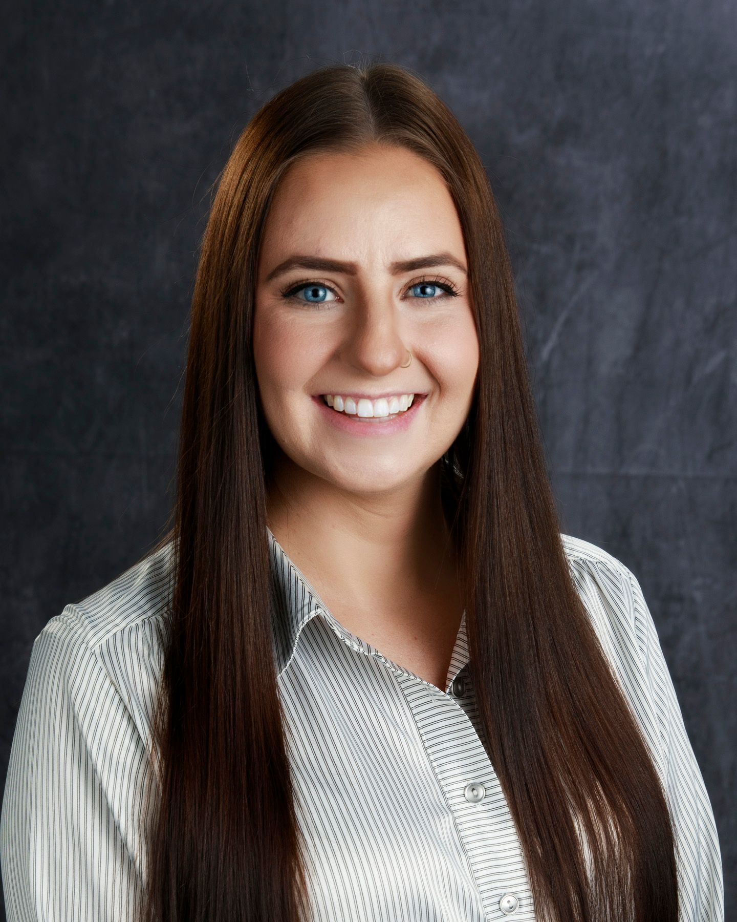 Young woman with long brown hair smiles, wearing a striped shirt against a dark backdrop.