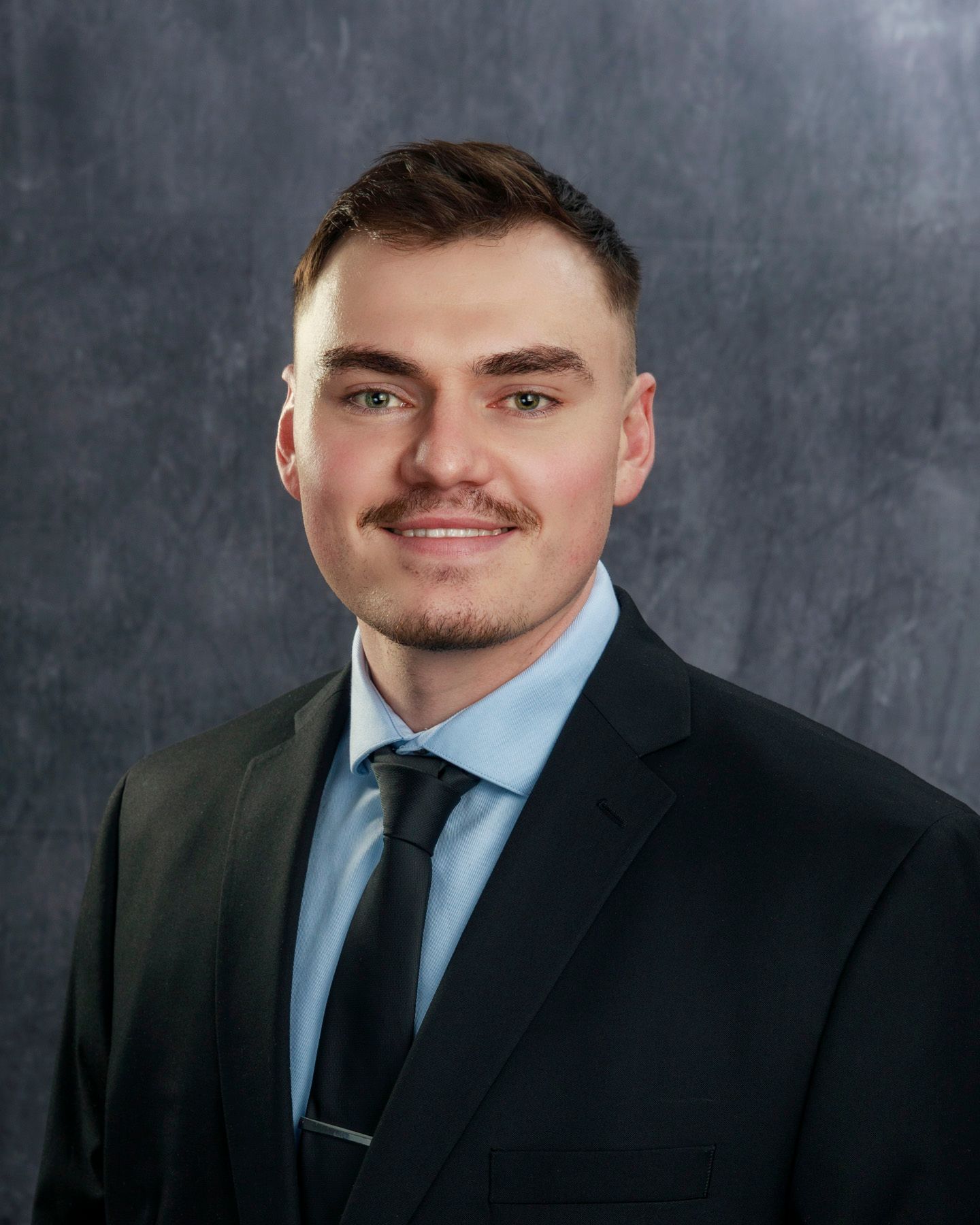 Man in a suit smiles at the camera, light blue shirt, black tie, dark hair and mustache, against a gray background.