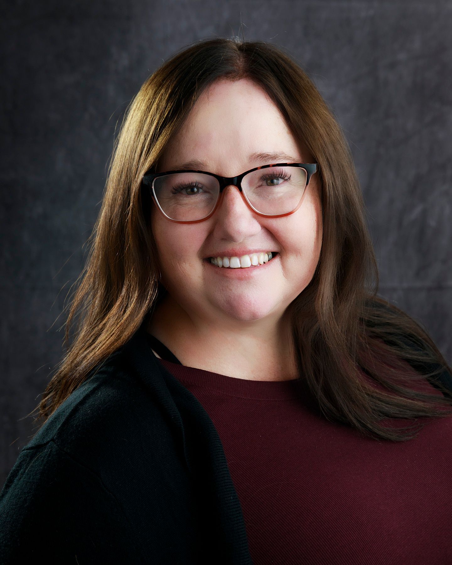 Woman with glasses smiles; maroon top and black cardigan, against a gray background.