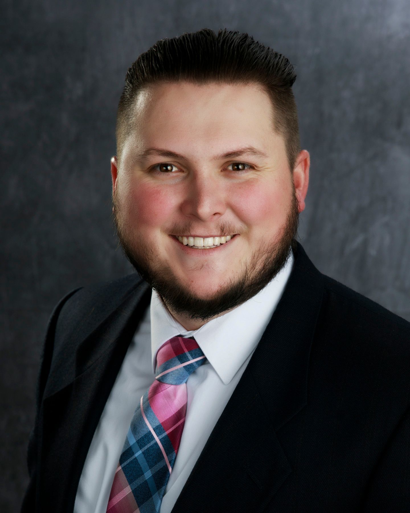 Man in suit with a pink and blue tie smiling at the camera, against a dark background.