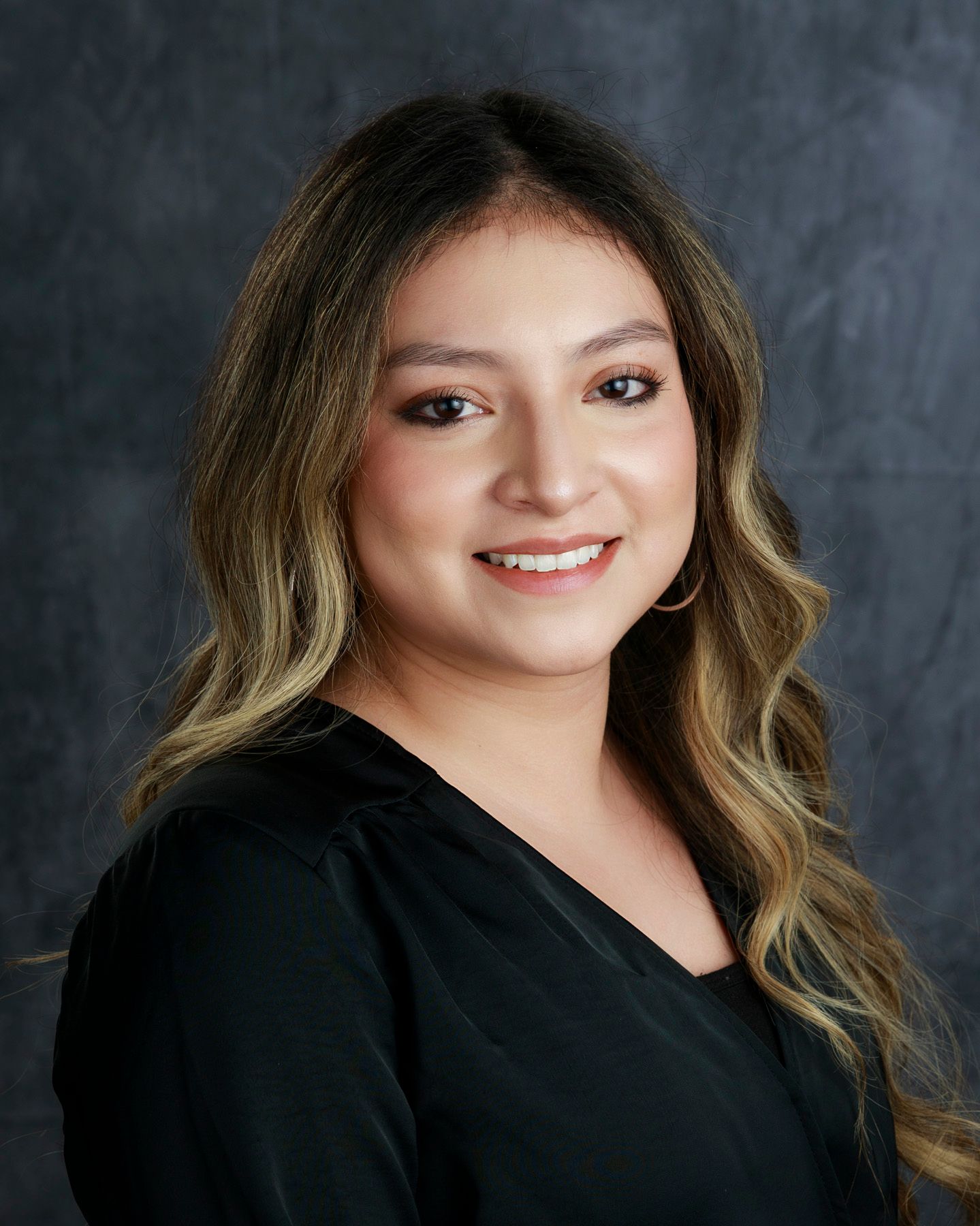 Woman with long, wavy brown hair, smiling, wearing a black top, and gold hoop earrings.