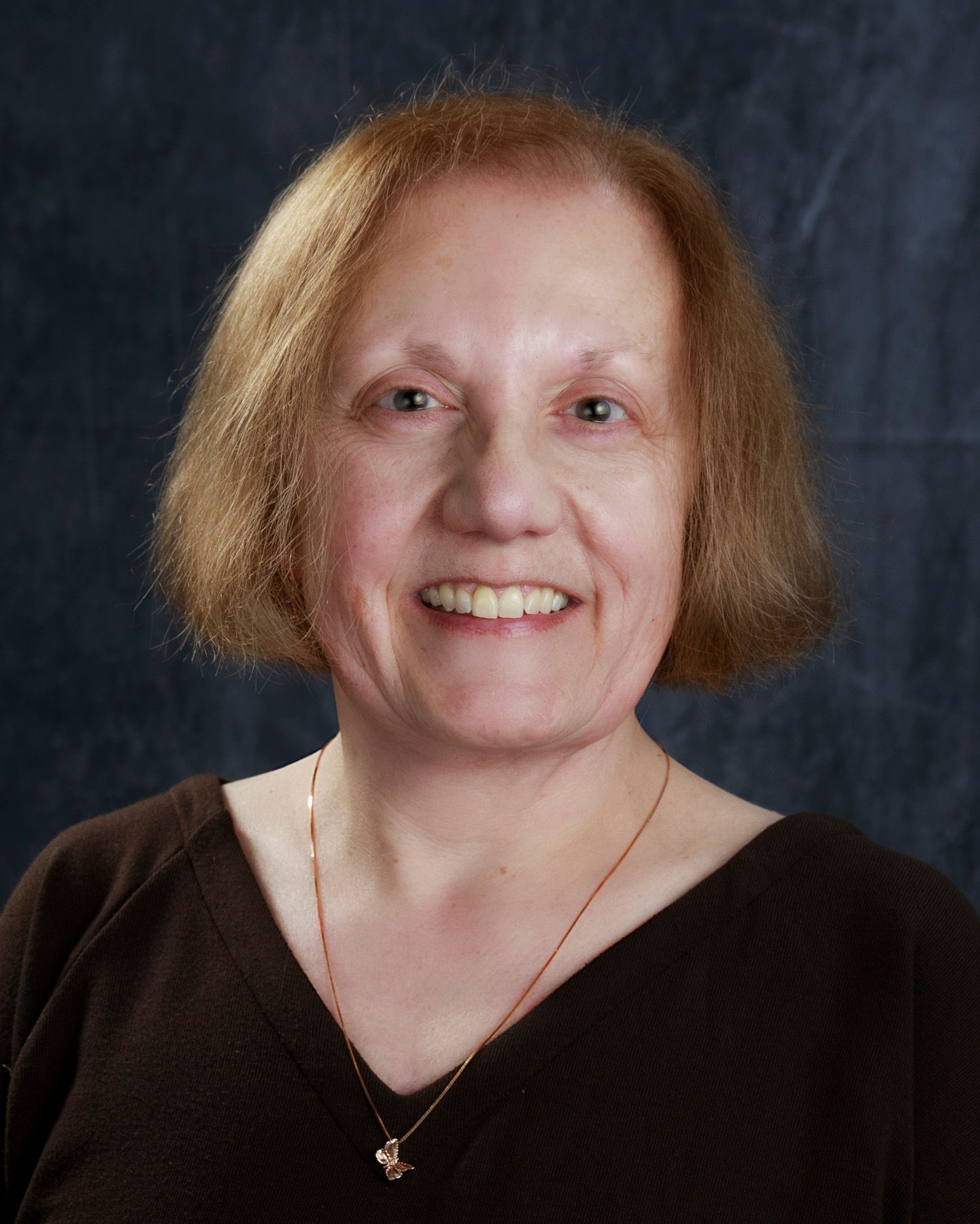 Woman with light brown hair smiles, wearing a brown shirt and a gold necklace, against a dark blue background.