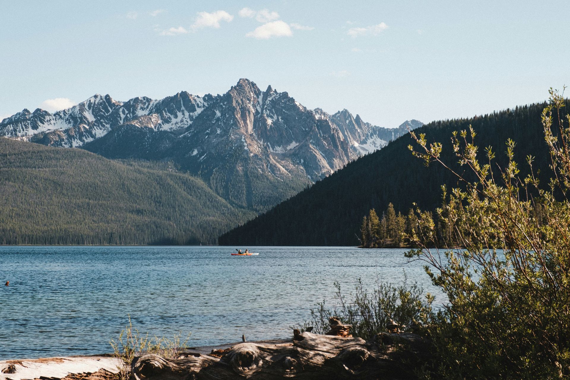 Lake with mountains in the background, blue water, and a person in a kayak. Sunny day with green trees.