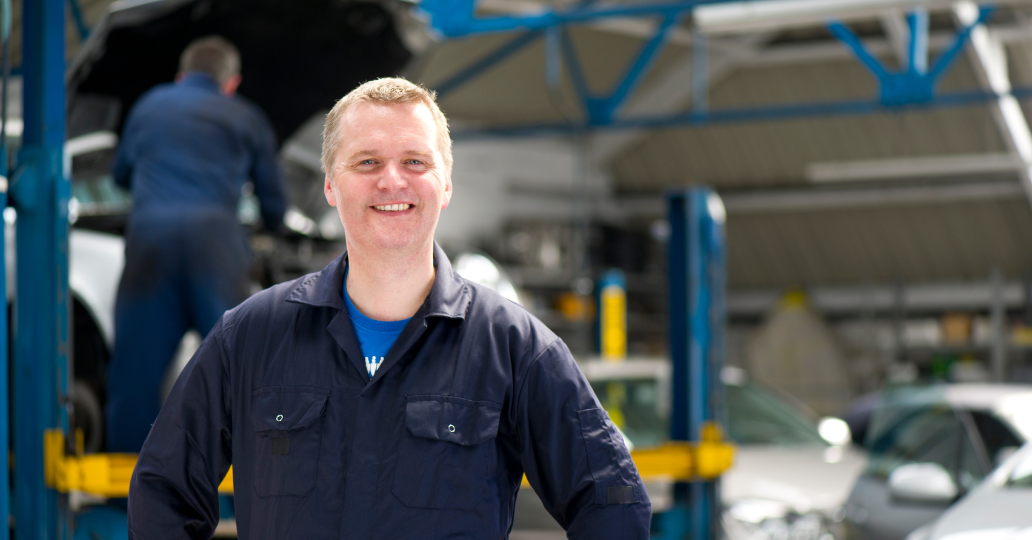 Smiling mechanic in blue jumpsuit, arms at side, in auto repair shop with another mechanic in background.