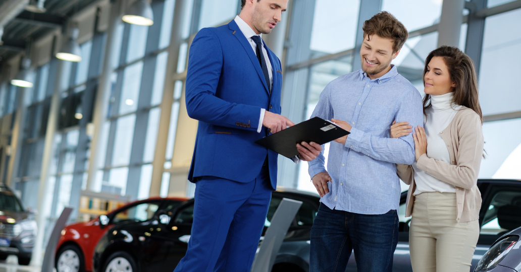 Car salesperson in blue suit shows documents to a smiling couple in a car showroom.