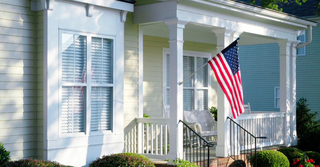 Cottage-style house with porch, American flag, white columns, and sunny exterior.