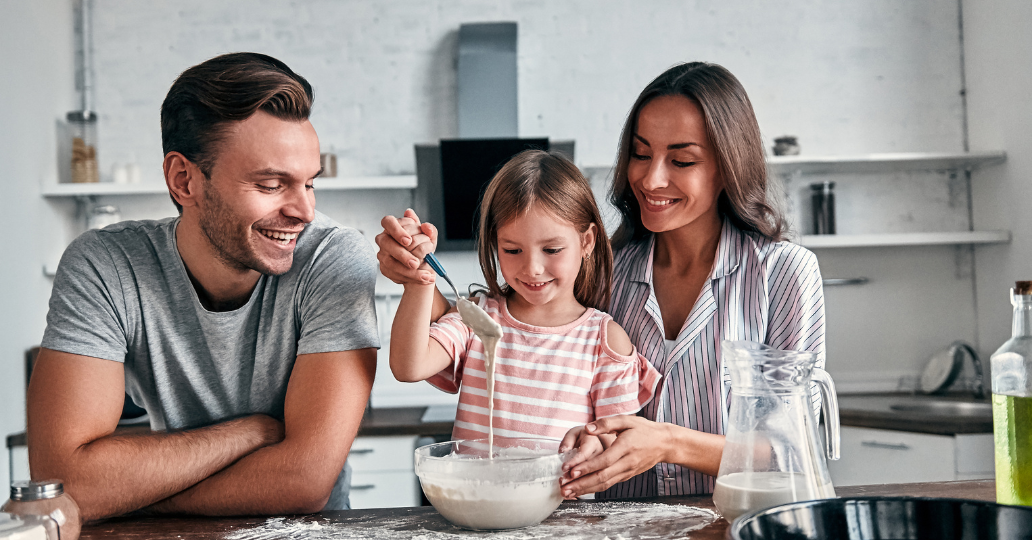 Family in kitchen, girl stirring batter, parents smiling.