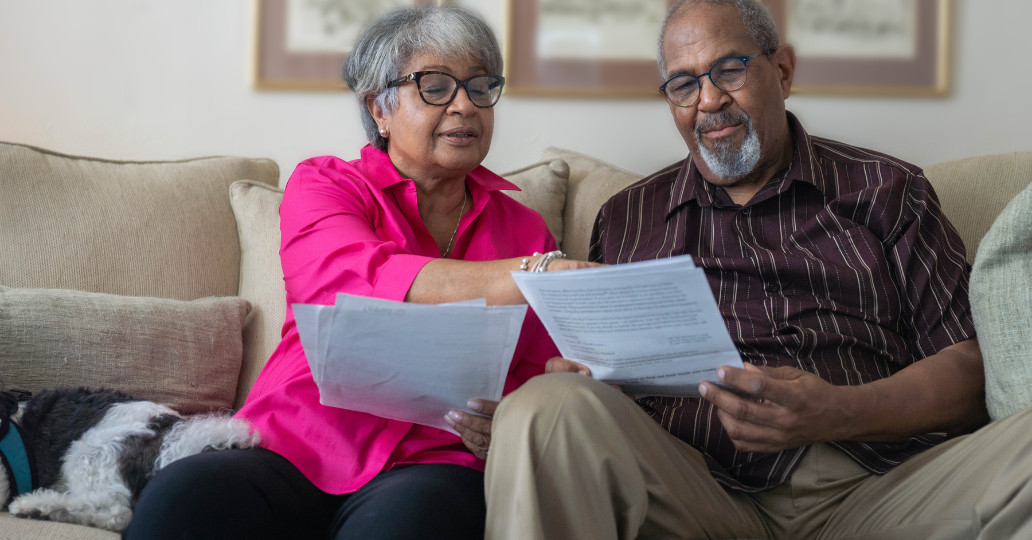 Couple on a couch reviewing papers; woman points, man smiles, dog nearby.