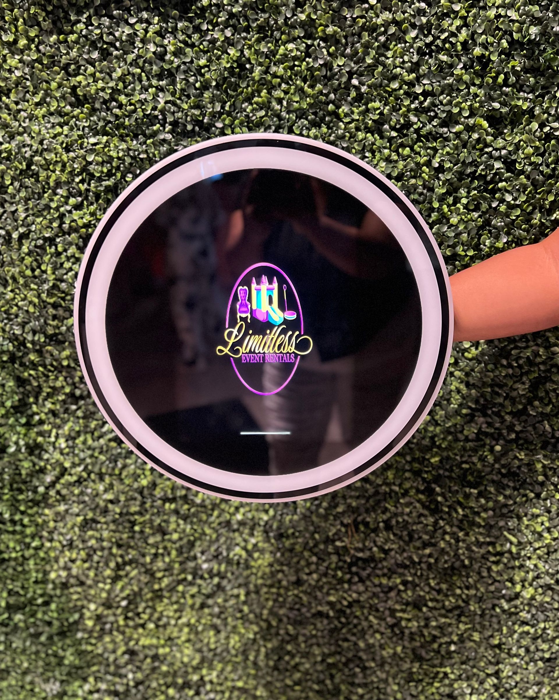 A person is holding a round mirror in front of a hedge wall.