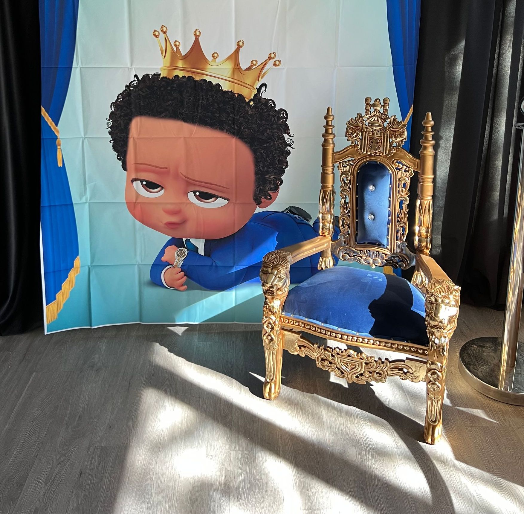 A baby in a crown is sitting in a chair in front of a boss baby backdrop.