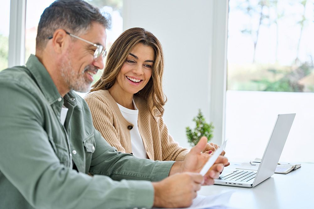 Man and woman smiling, looking at papers and laptop. White table, bright room.