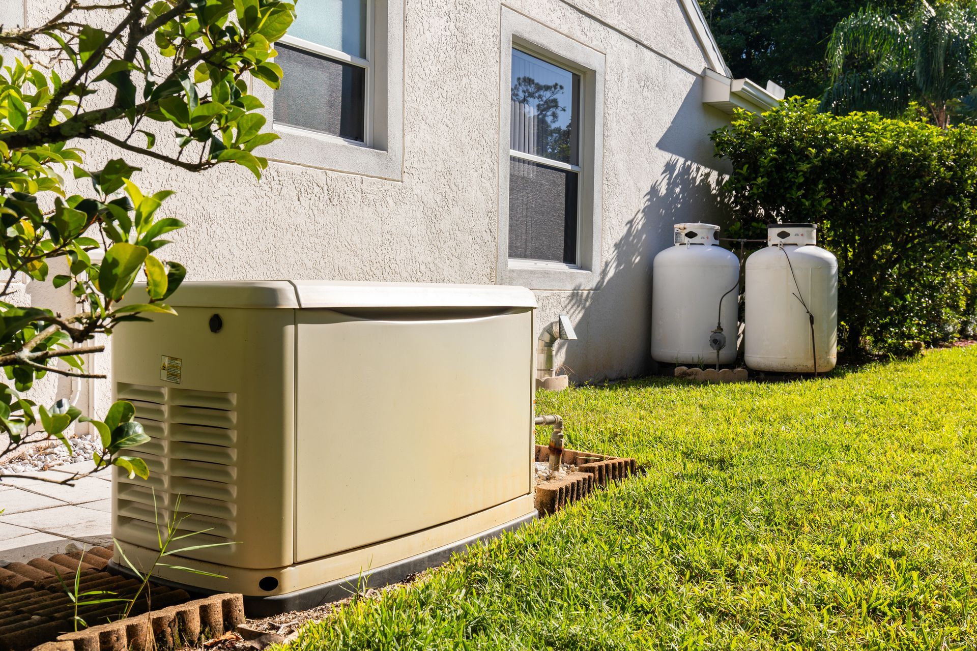 Backup generator on concrete pad beside house, with propane tanks and greenery nearby.