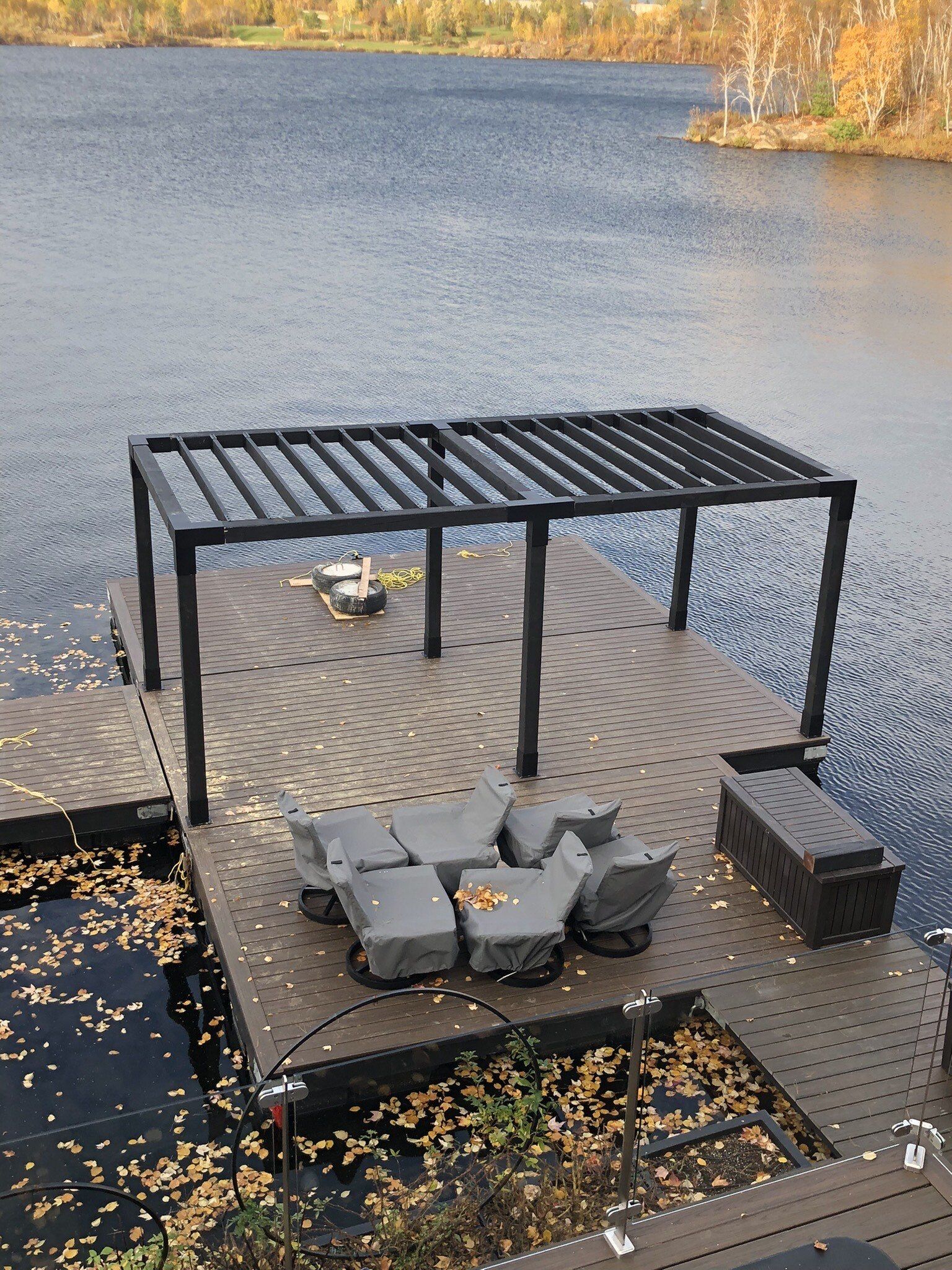 A dock with a pergola and chairs on it overlooking a lake.