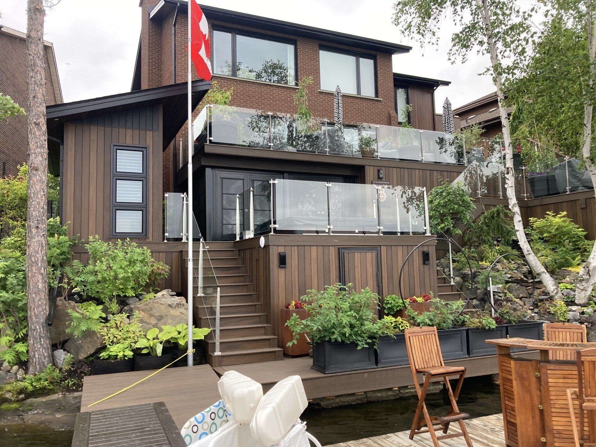 Brown lakeside house with multiple levels, a deck, glass railings, and a Canadian flag.