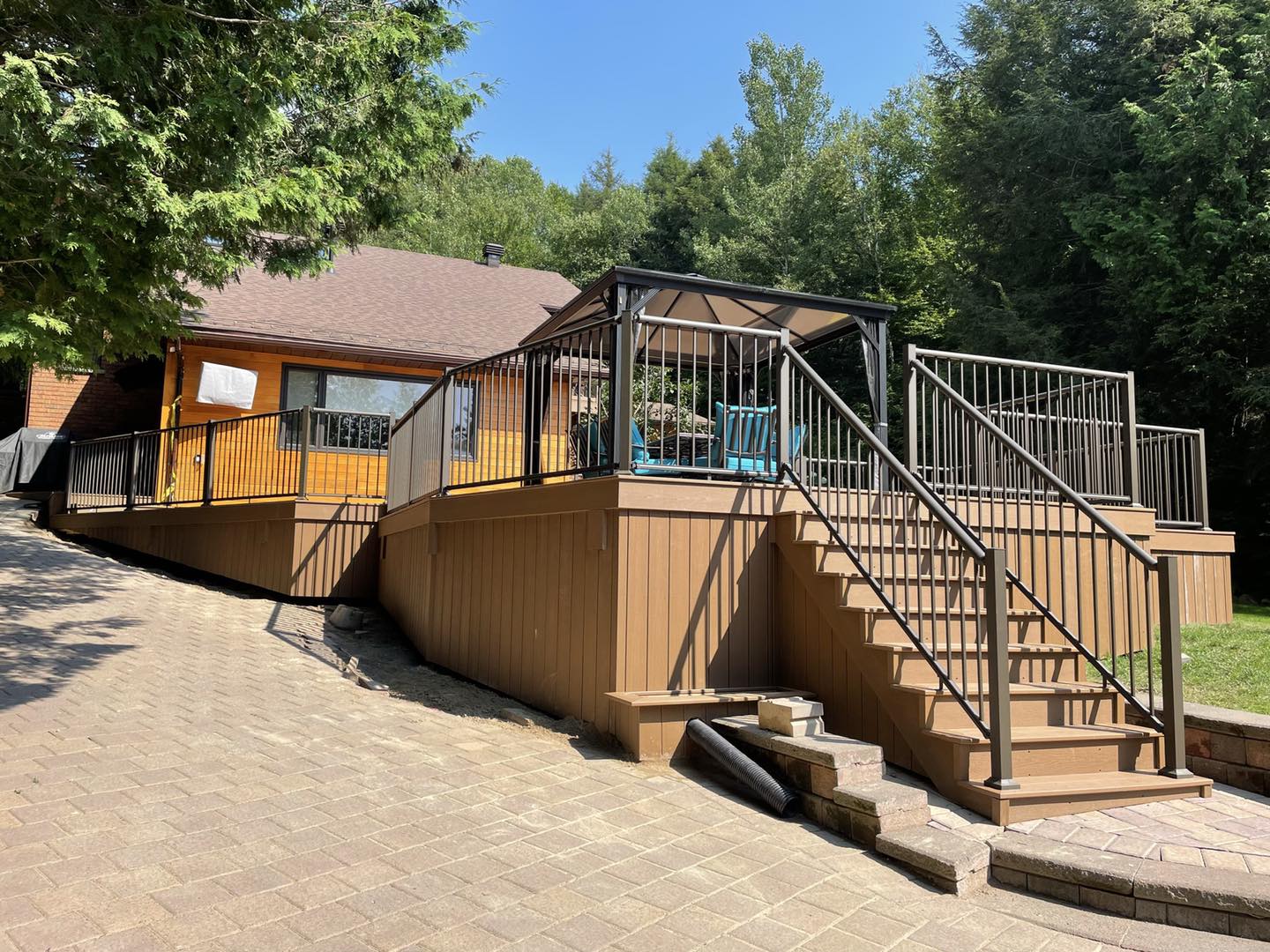 Brown deck and house with stairs, gazebo, and surrounding trees. Paved patio in front.