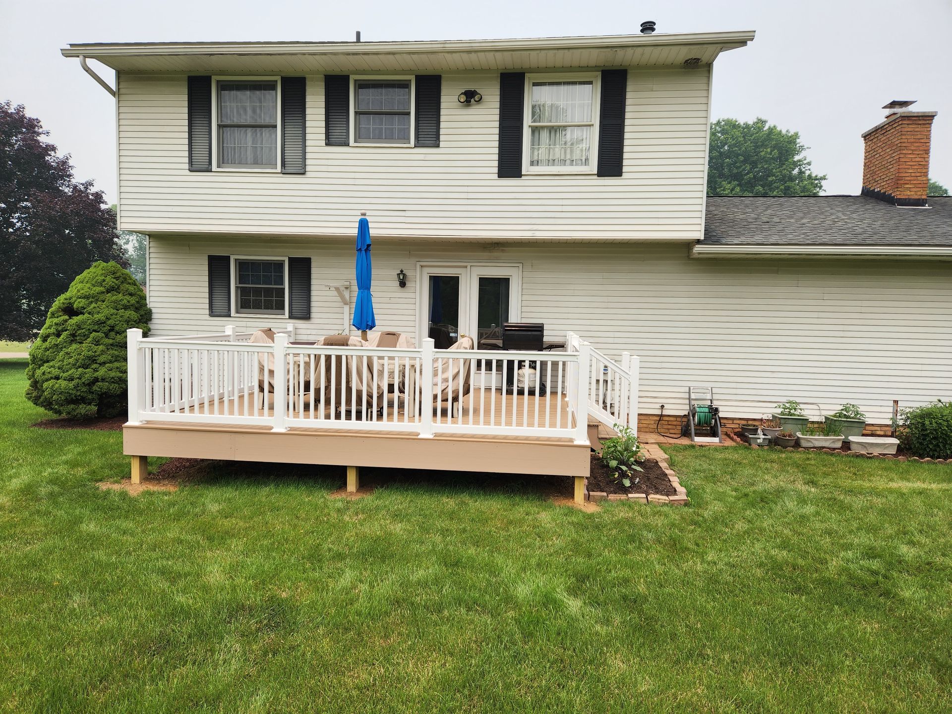 A two-story white house with a deck, outdoor furniture, blue umbrella, and a large lawn under a cloudy sky.