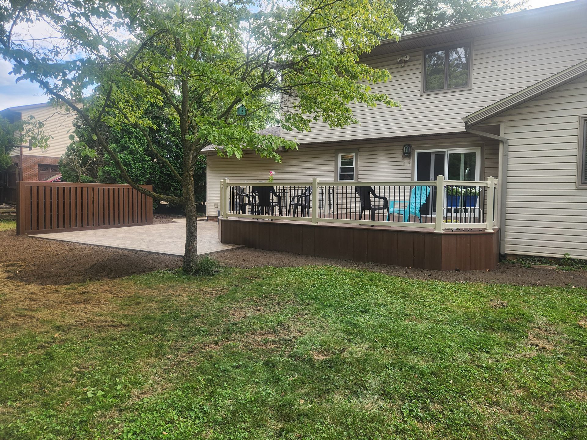 A house with a backyard featuring a brown wooden deck, a matching privacy fence, and a patio area under a large tree.
