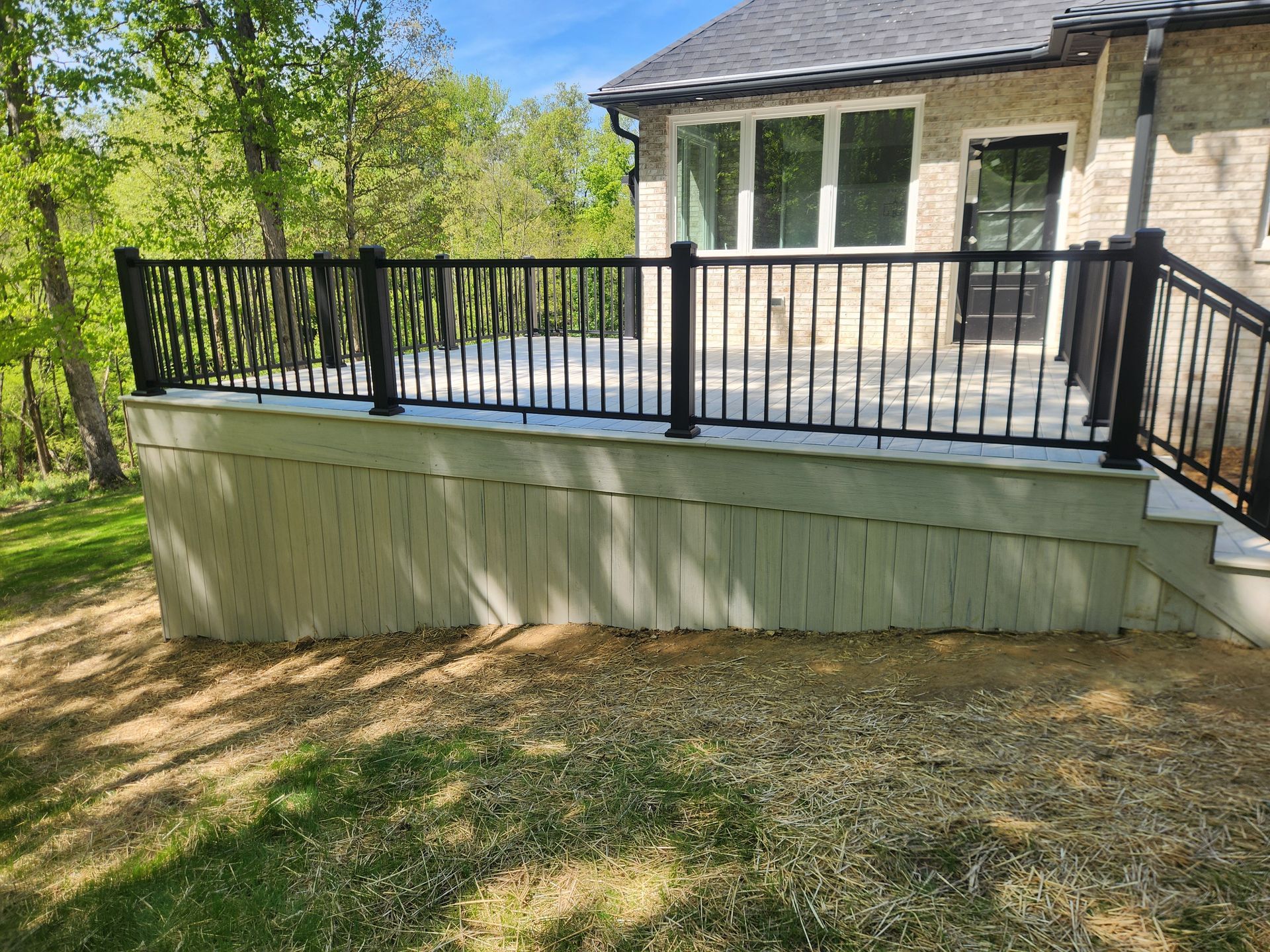 A stone house with a light-colored patio deck featuring black metal railings, surrounded by trees and a dirt yard.
