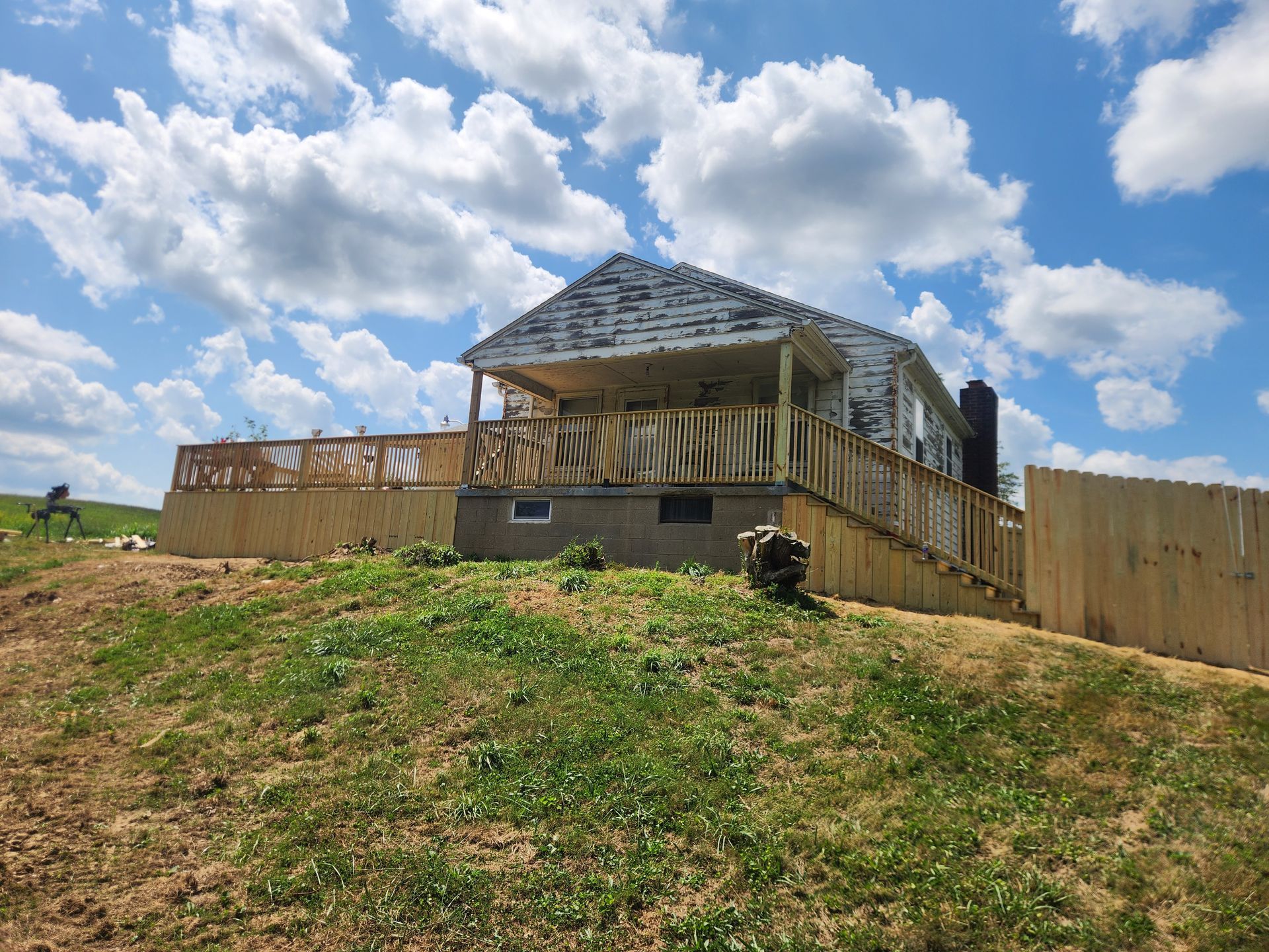 A house with peeling paint and a large, newly constructed wooden deck and fence on a grassy hill under a cloudy blue sky.