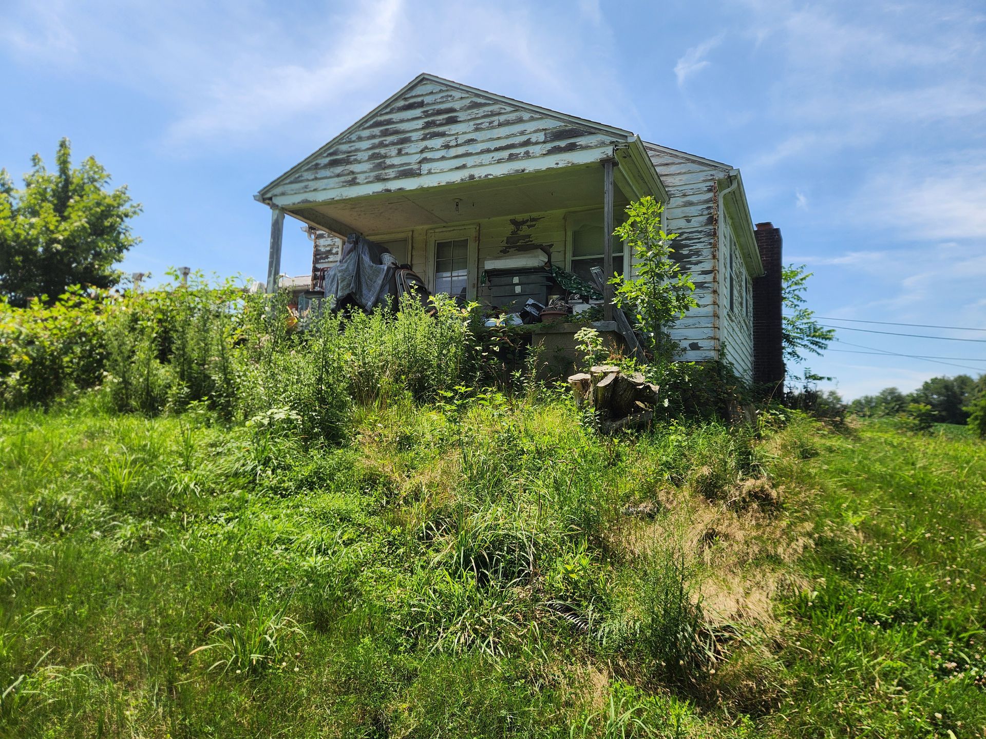 A small, weathered white cottage with a porch sits on a grassy hill under a blue sky, surrounded by overgrown vegetation.