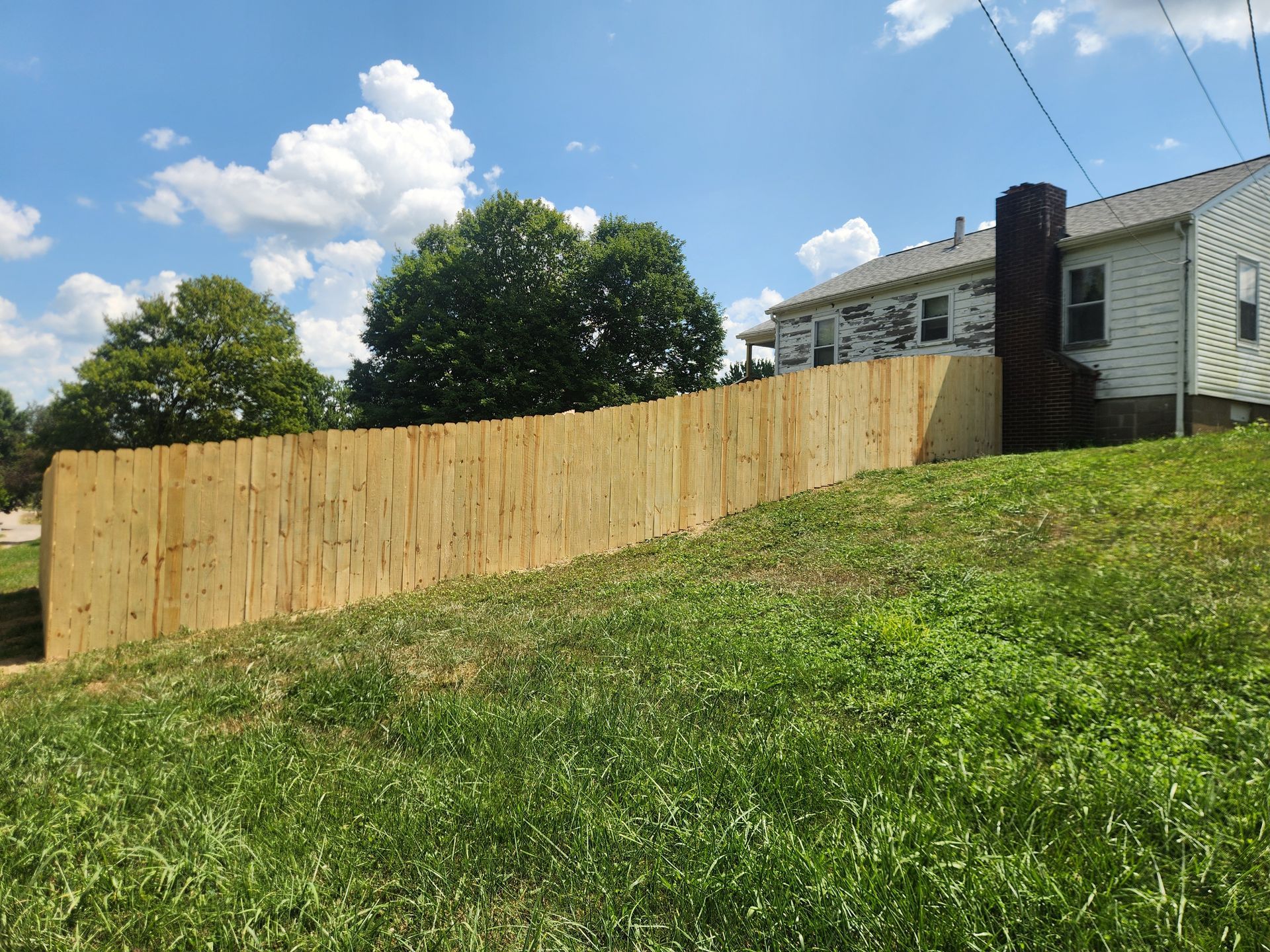 A new wooden fence runs along a grassy slope next to a house with white siding and a dark chimney under a blue sky.