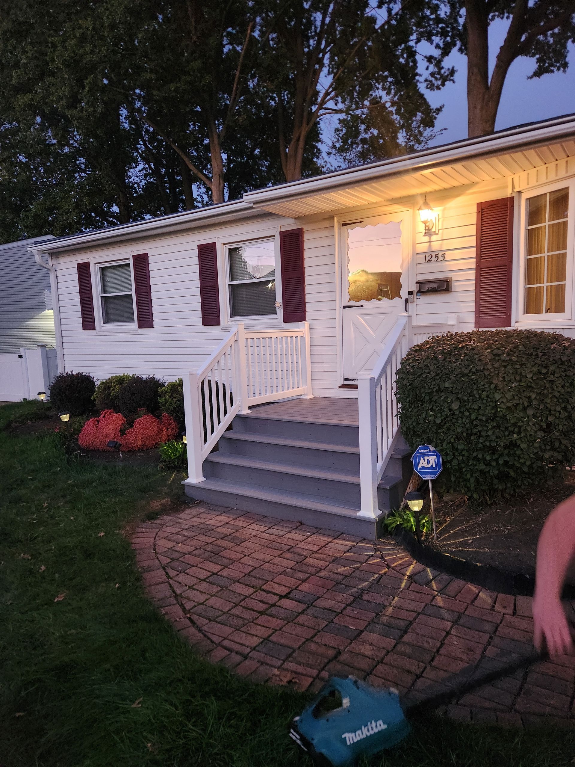 A teal leaf blower held at the edge of a brick walkway in front of a white house with maroon shutters and gray porch steps.