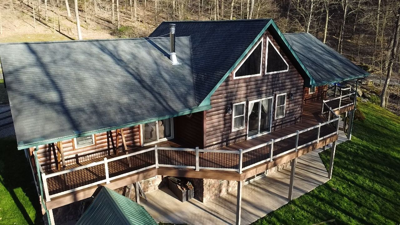 An aerial view of a rustic, multi-level wooden cabin with a dark shingled roof, large deck, and surrounding forest.