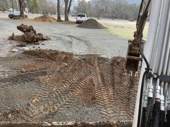 An excavator digging in a gravel area, with a pile of dirt and vehicles in the background.