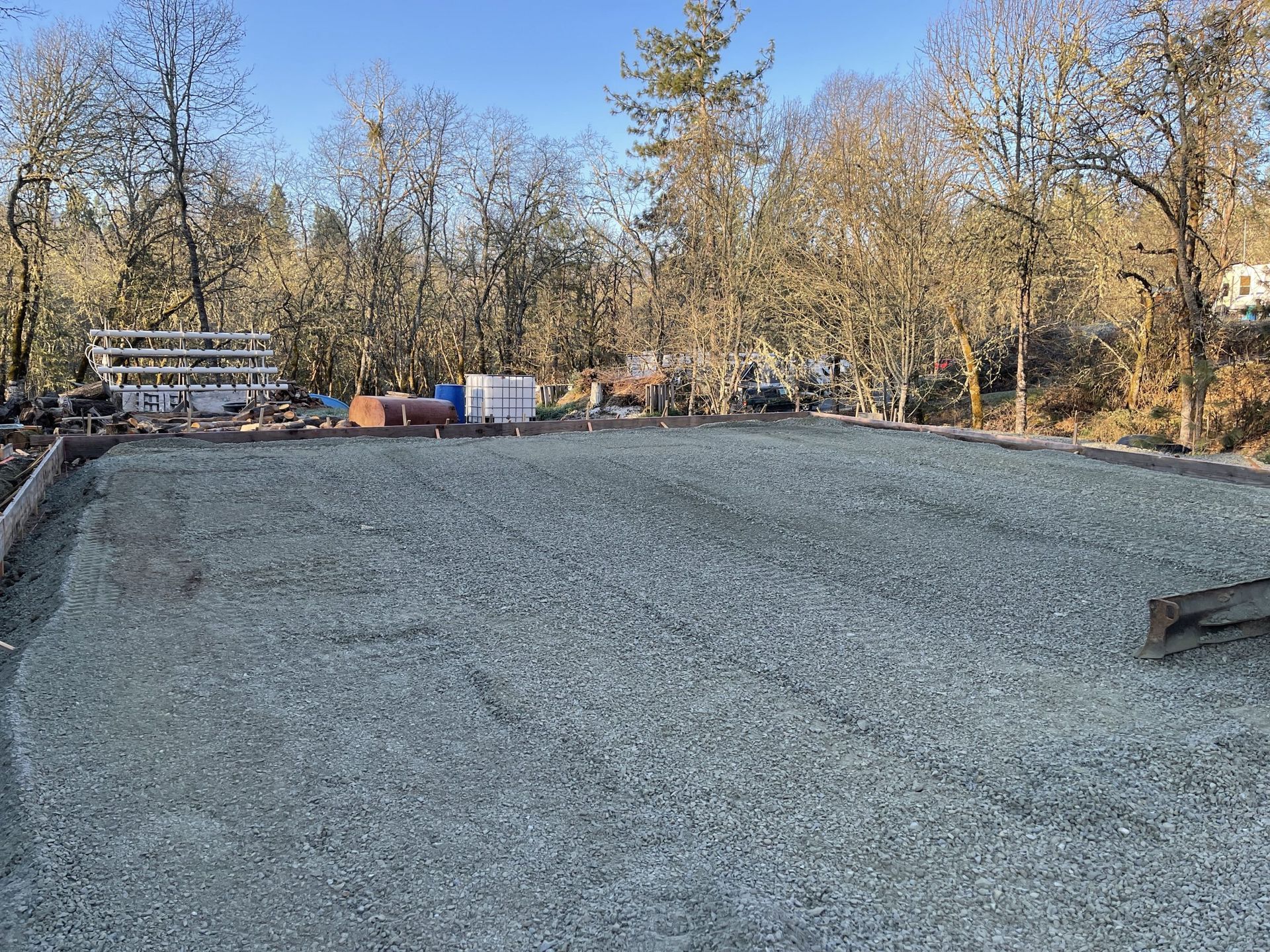 Gravel-covered ground prepared for construction, construction materials in the background, trees under a blue sky.