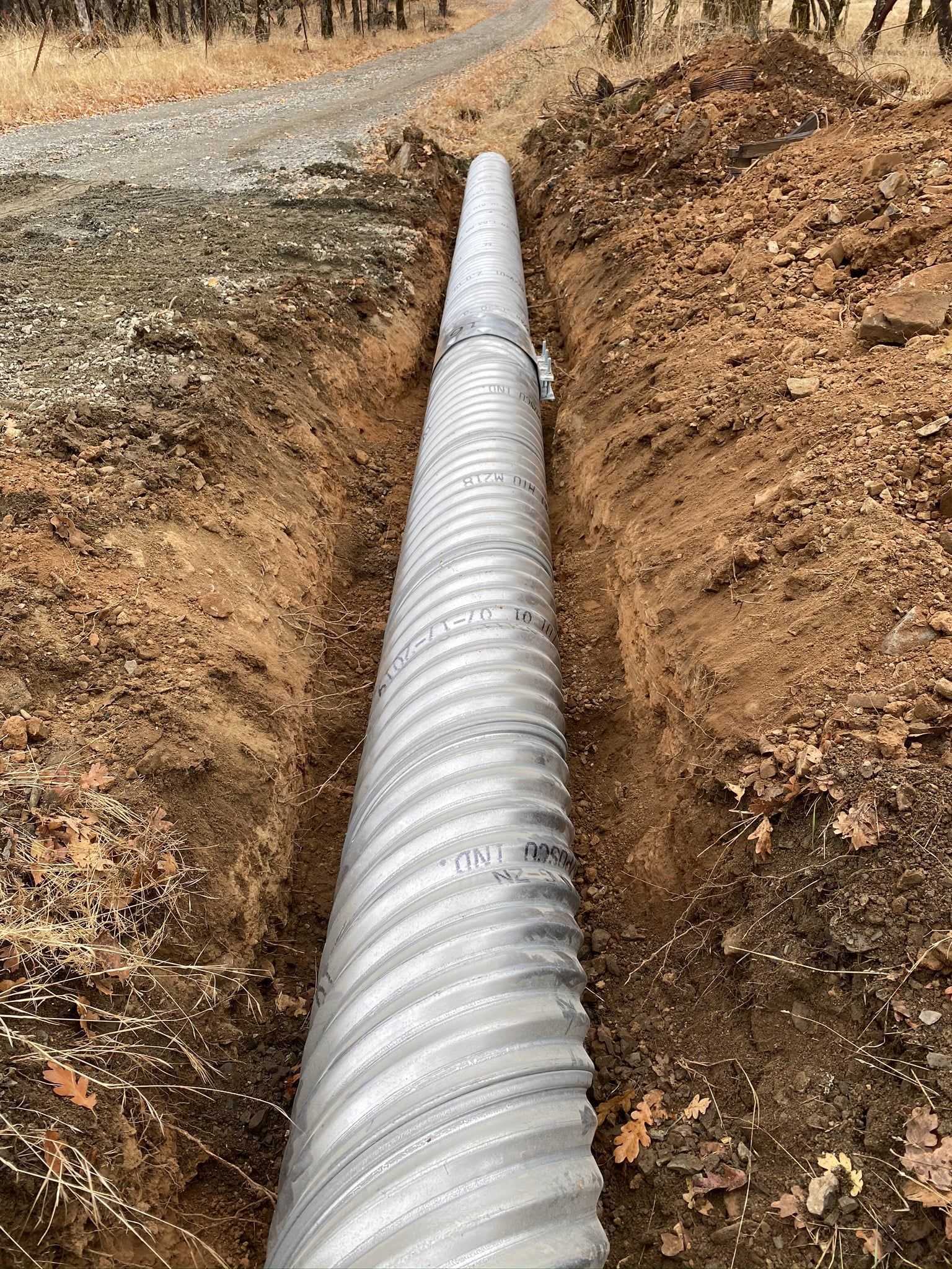 Corrugated metal pipe in a trench, alongside a gravel road, in a dirt setting.