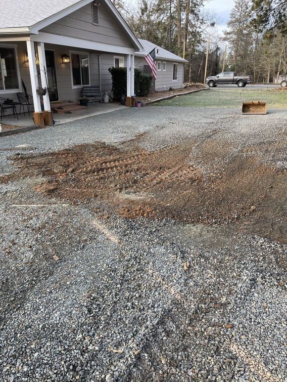 Gravel driveway with disturbed dirt patch in front of a small gray house with a porch and an American flag.
