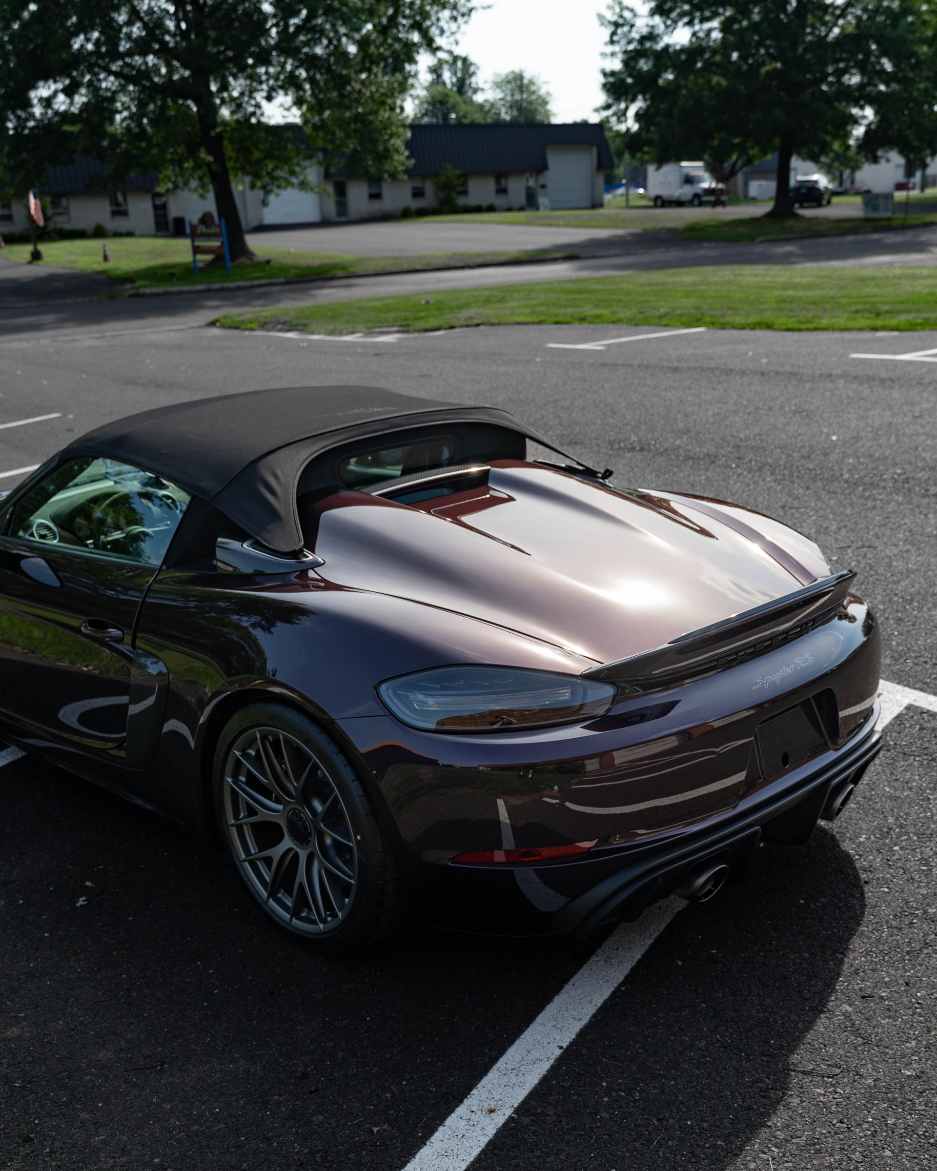 Dark convertible sports car parked in an asphalt lot. Black soft top, metallic paint, dark wheels, trees in the background.