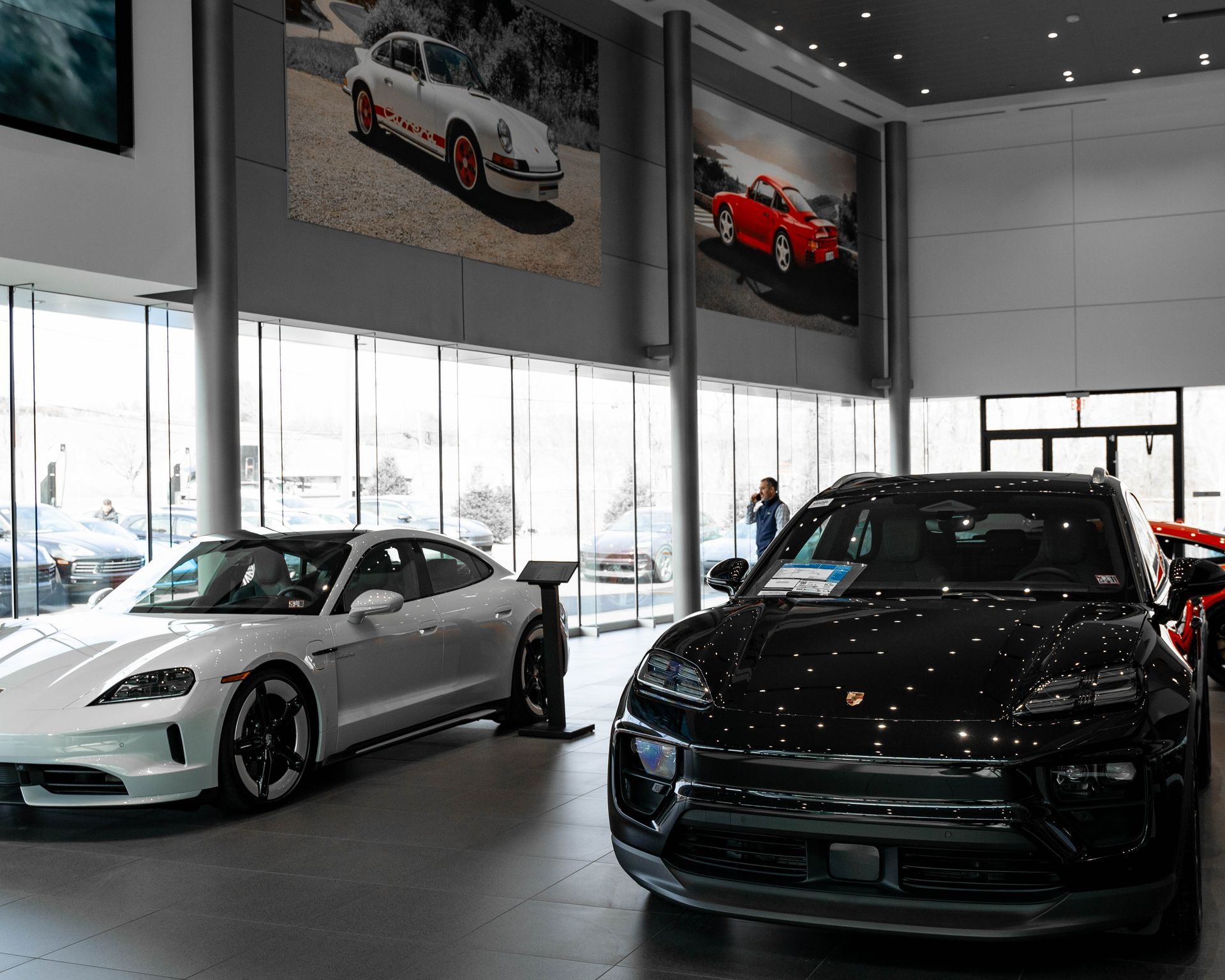 Inside a Porsche dealership, a white Taycan and a black Macan SUV are on display; photo prints of classic Porsches hang on the wall.