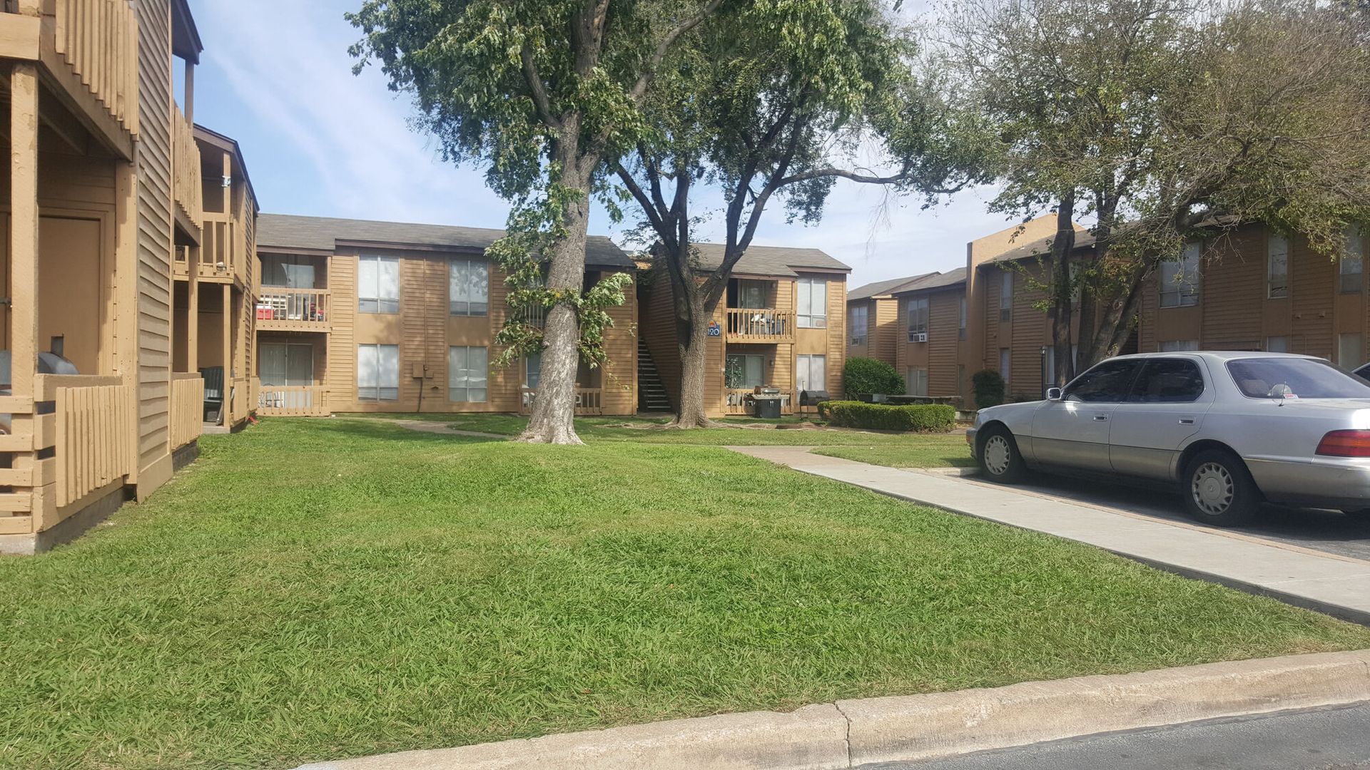 Apartment complex exterior with a grassy lawn, trees, and a parked silver car.