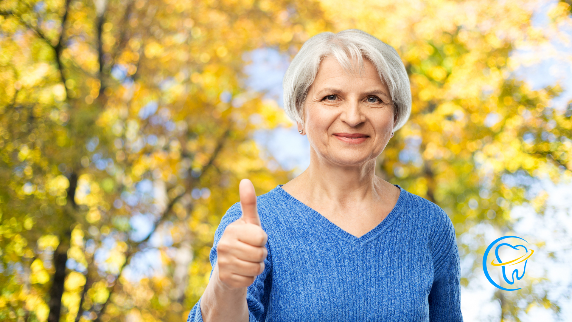 An elderly woman in a blue sweater is giving a thumbs up.