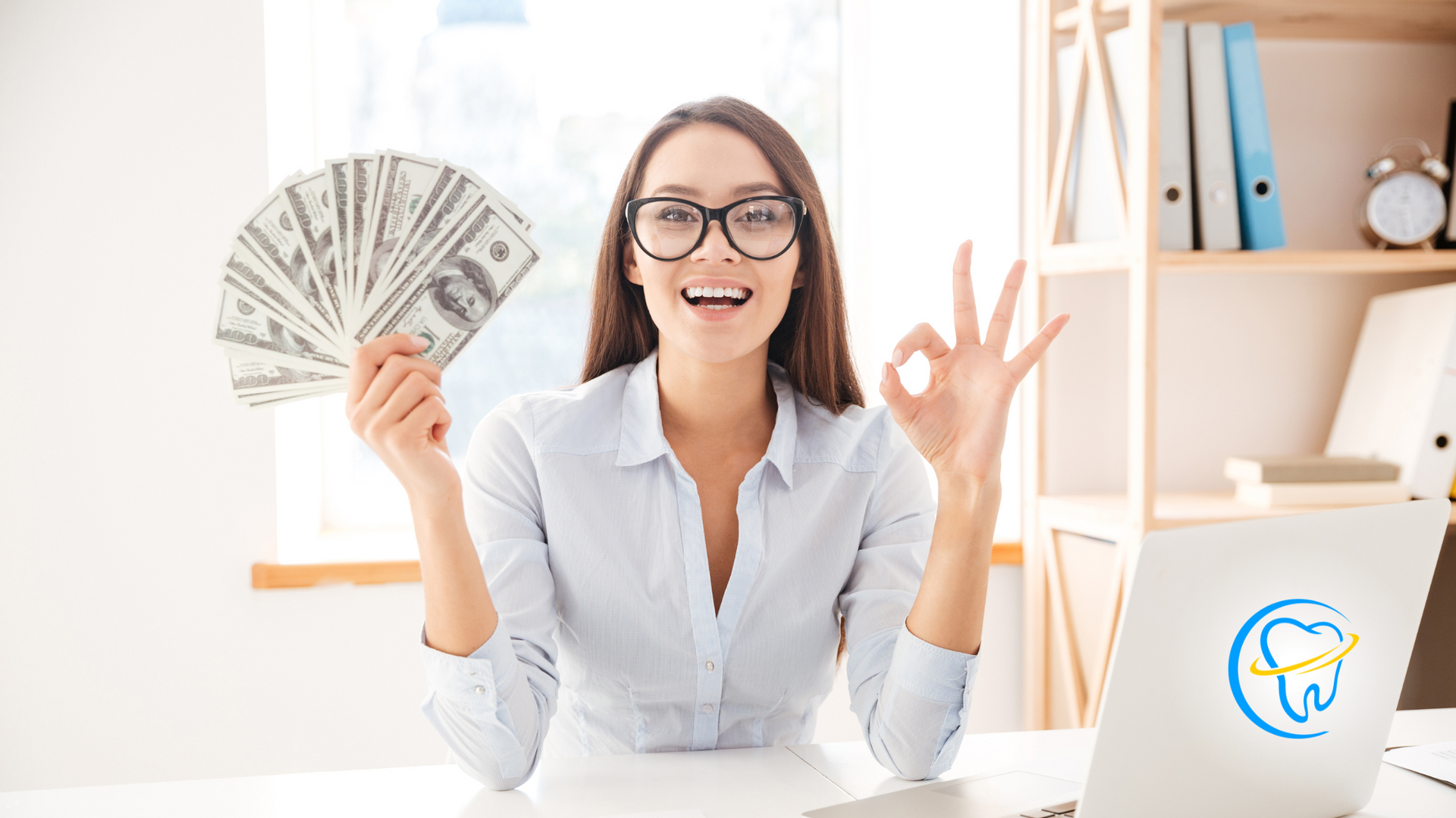 A woman is sitting at a desk holding a fan of money and giving an ok sign.