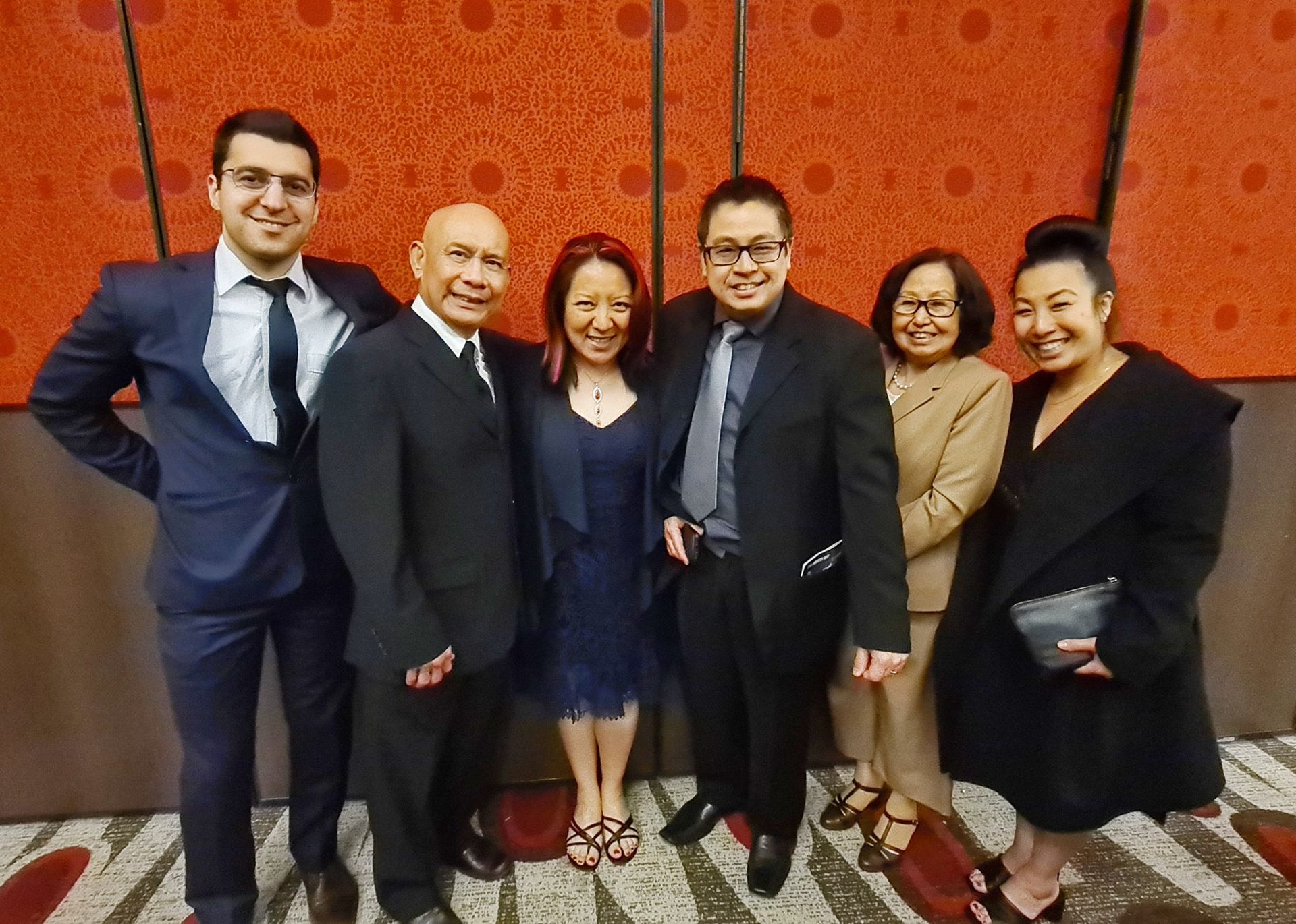 Group of seven people posing in front of a red textured wall. Men in suits, women in dresses.