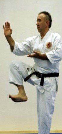 A man in a white karate uniform with a black belt is standing on one leg.