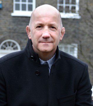 A bald man wearing a black coat and tie is standing in front of a building.