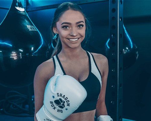 A woman is wearing boxing gloves and smiling in a gym.