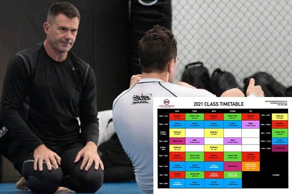 A man is kneeling next to a man sitting on a mat next to a class timetable