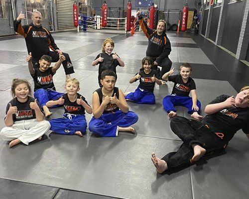 A group of children are sitting on a mat in a gym giving thumbs up.