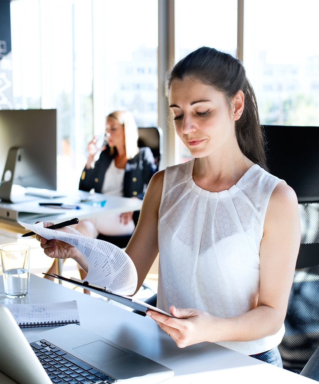 A woman is sitting at a desk in front of a laptop computer holding a piece of paper.
