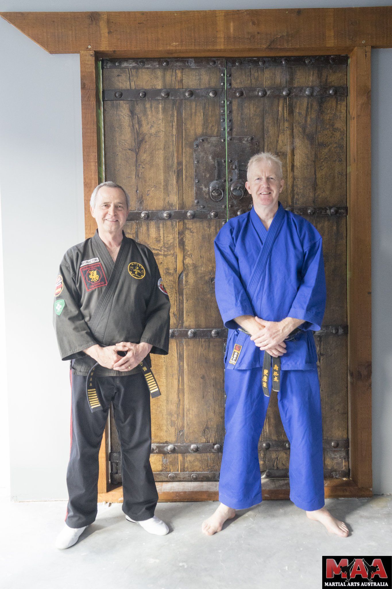 Two men in karate uniforms are standing next to each other in front of a wooden door.